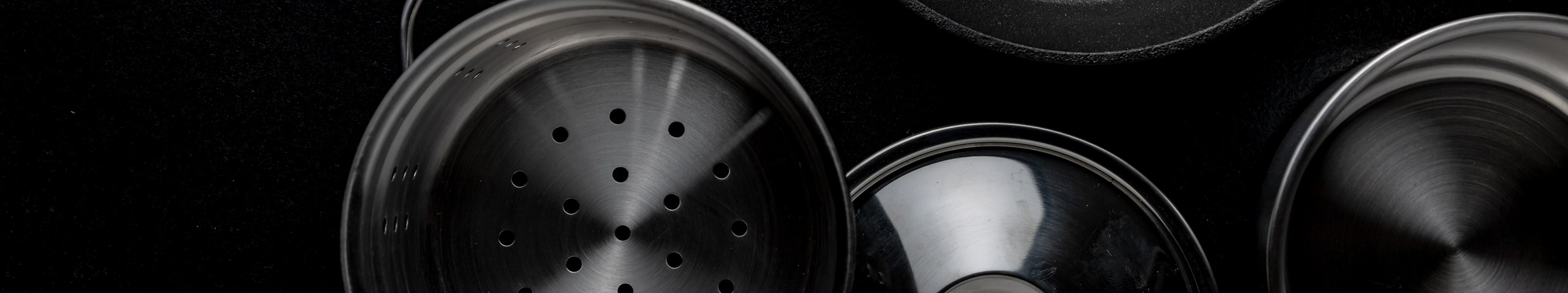 Pentole, padella in ghisa, cestello forato e coperchi in acciaio inox visti dall alto su fondo nero