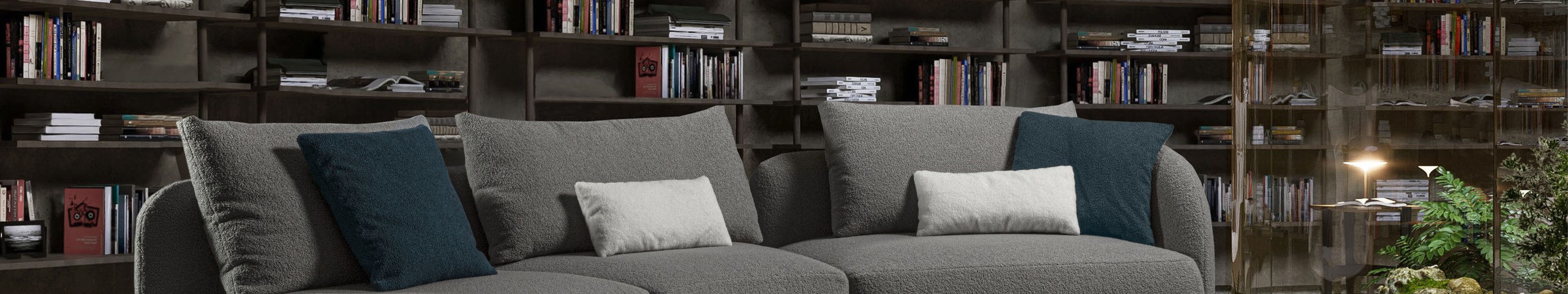 Divano modulare curvo rivestito in tessuto bouclé grigio con cuscini blu petrolio, in una sala con libreria a parete