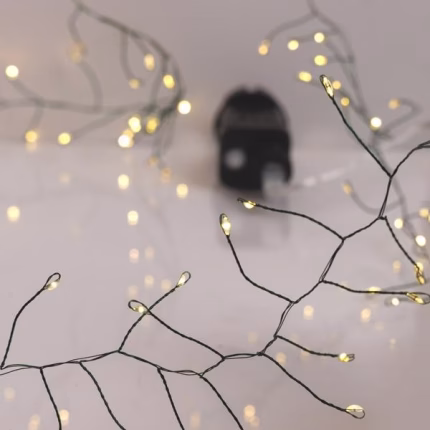Catena luminosa Cluster accesa: mini LED bianco caldo ravvicinati su cavo verde ramificato, con l’alimentatore nero appoggiato su un fondo chiaro