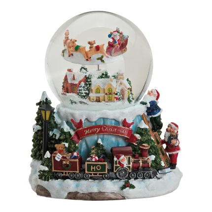 Carillon natalizio su fondo bianco: base decorata con una stazione ferroviaria innevata, un treno, Babbo Natale, bambini e abeti; sopra, una sfera di vetro con la neve e una slitta trainata dalle renne.