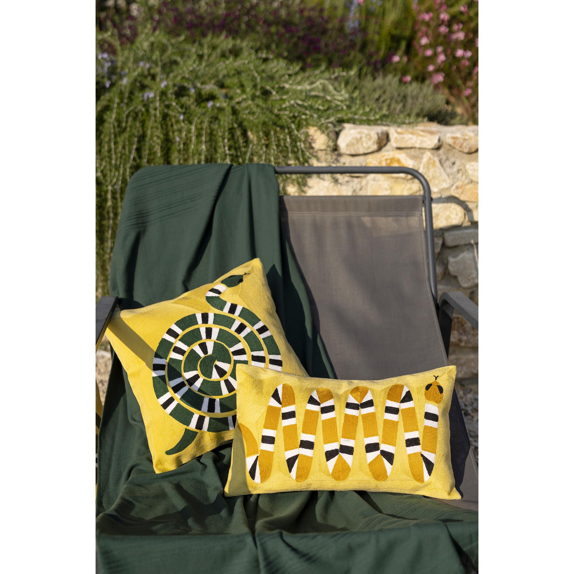 Due cuscini Yoka giallo mango, il quadrato 45 × 45 cm e il rettangolare 30 × 50 cm, appoggiati su un plaid verde scuro steso su una sdraio da giardino davanti a un muretto in pietra fiorito. Fotografia di catalogo del marchio Vivaraise.
