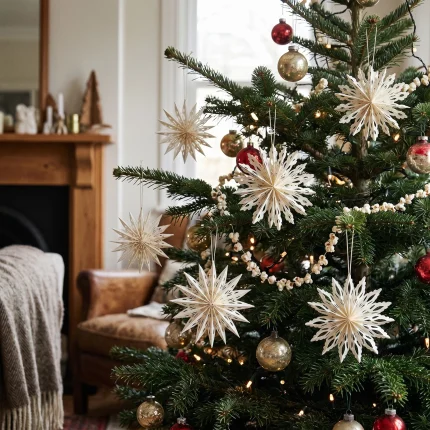 I fiocchi di neve in carta bianca appesi ai rami di un albero di Natale decorato, accanto a palline dorate, davanti a una finestra e a una poltrona con plaid. Albero, palline, poltrona, plaid e arredi non sono compresi.