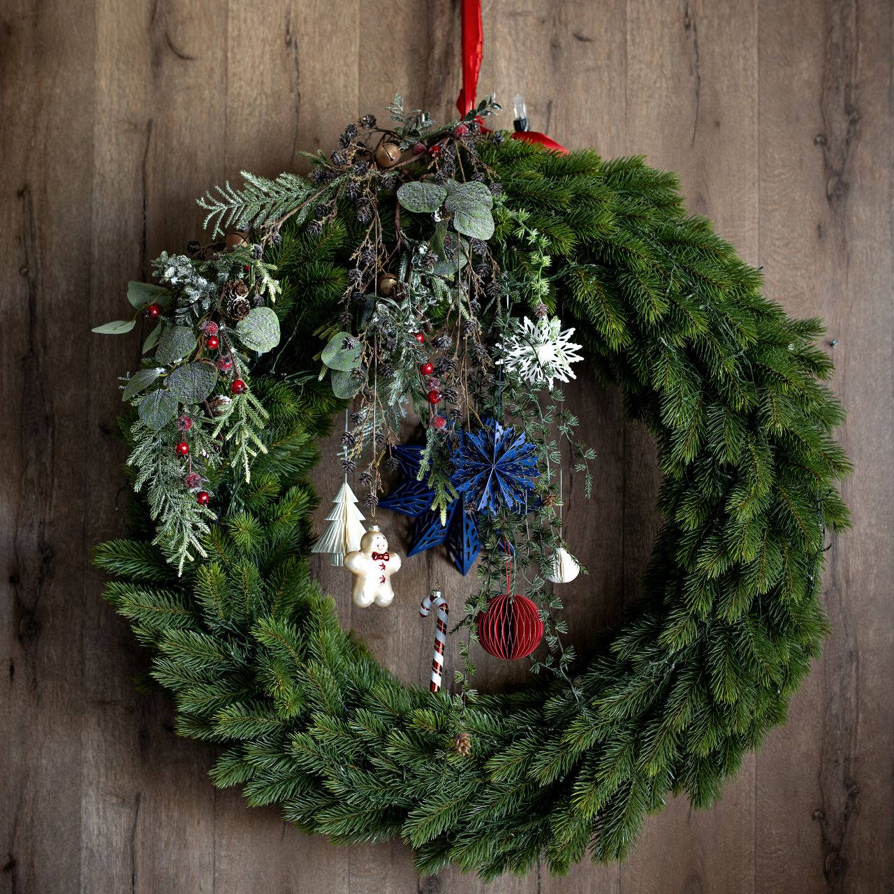 I fiocchi di neve in carta bianca inseriti fra i rami di una ghirlanda di abete appesa a una parete di legno, insieme a fiori secchi azzurri e bacche. Ghirlanda, fiori, bacche e parete non sono compresi.