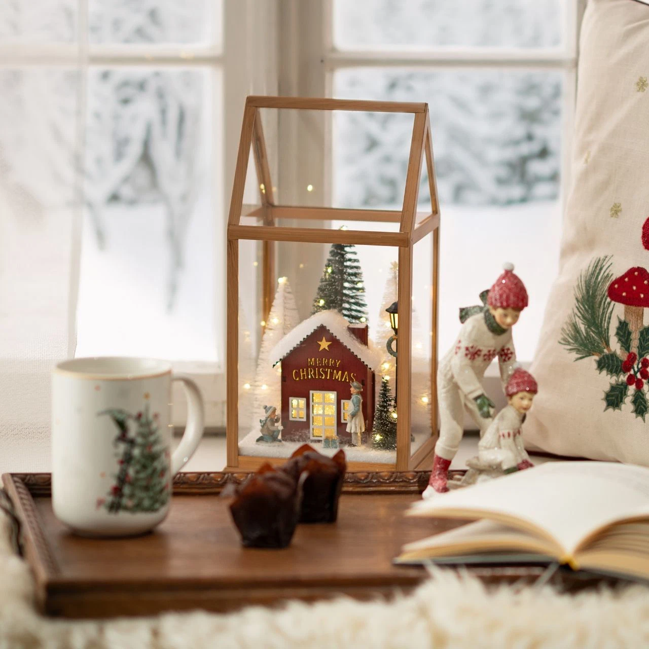 La lanterna color legno accesa su un vassoio accanto a una finestra innevata, con una tazza bianca decorata, alcuni dolcetti, un libro aperto, un cuscino natalizio e un plaid. Vassoio, tazza, dolcetti, libro, cuscino, plaid e arredi non sono compresi.