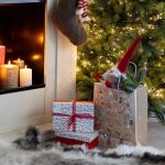 Il sacchetto regalo in carta kraft appoggiato davanti a un camino acceso accanto a un albero di Natale illuminato, con dentro un folletto di Natale; davanti un pacco regalo bianco e rosso, sopra una calza della Befana appesa e candele accese sulla mensola. Folletto, pacco regalo, calza, candele, albero, camino e tappeto non sono compresi.