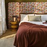 Camera da letto con testiera e cuscini nel tessuto Nira di Villa Nova, colorazione Plume, copriletto marrone, comodino con lampada e vaso. In vendita c’è soltanto il tessuto al metro: arredi e complementi della scena non sono compresi.
