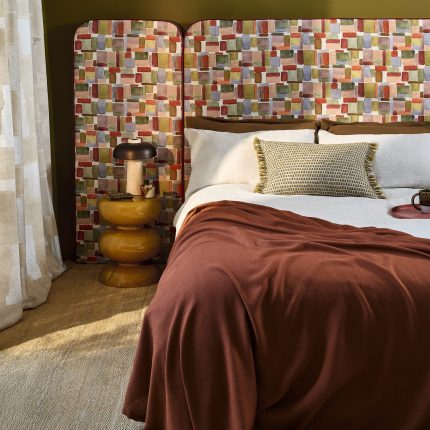 Camera da letto con testiera e cuscini nel tessuto Nira di Villa Nova, colorazione Plume, copriletto marrone, comodino con lampada e vaso. In vendita c’è soltanto il tessuto al metro: arredi e complementi della scena non sono compresi.