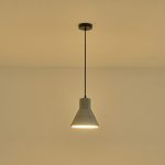 Lampada a sospensione Arme in cemento grigio sfaccettato, diametro 20 cm, accesa contro una parete chiara. La lampadina non è compresa