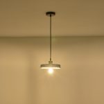 Lampada a sospensione Yeso in cemento grigio a disco, diametro 21 cm, accesa contro una parete chiara. La lampadina non è compresa