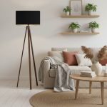 Lampada da terra a treppiede in legno noce con paralume nero accanto a un divano in soggiorno; divano, tavolino, tappeto, piante e oggetti della scena non sono compresi