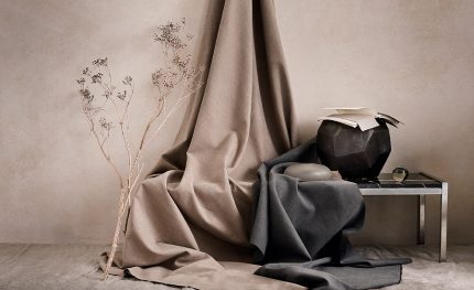 Tessuto Linara di Romo nei toni naturali del beige e del grigio, drappeggiato su una seduta accanto a rami secchi e a un vaso scuro. In vendita c’è soltanto il tessuto al metro: arredi e complementi della scena non sono compresi.