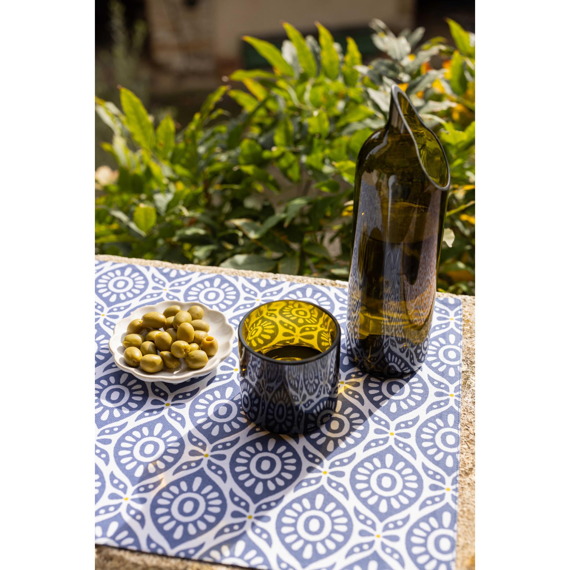 Tovaglietta Citrus Cobalt su un tavolo in giardino con bottiglia, bicchiere e olive; gli altri oggetti della scena non sono compresi