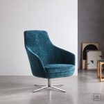 Poltroncina Kea a schienale medio rivestita in velluto blu, con basamento girevole a quattro razze in metallo cromato, in un soggiorno dalle pareti chiare e dal pavimento in cemento; accanto si vedono un tavolino tondo con piano in vetro e gambe in legno e due stampe astratte incorniciate appoggiate alla parete. Ambientazione generata con AI a partire dalla fotografia del prodotto: il tavolino, le stampe e il resto dell'allestimento non sono compresi.