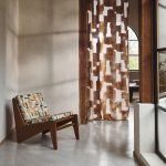 Poltroncina in legno con cuscini di seduta e schienale nel tessuto Nira di Villa Nova, colorazione Sienna, accanto a una tenda a tutta altezza in velo Idris Sienna, in un soggiorno con pavimento in cemento. In vendita c’è soltanto il tessuto al metro: arredi e complementi della scena non sono compresi.