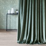 Tenda a tutta altezza in velluto Barcelona di Villa Nova, colorazione Spruce, davanti a una parete chiara con consolle nera e vasi bianchi. In vendita c’è soltanto il tessuto al metro: arredi e complementi della scena non sono compresi.