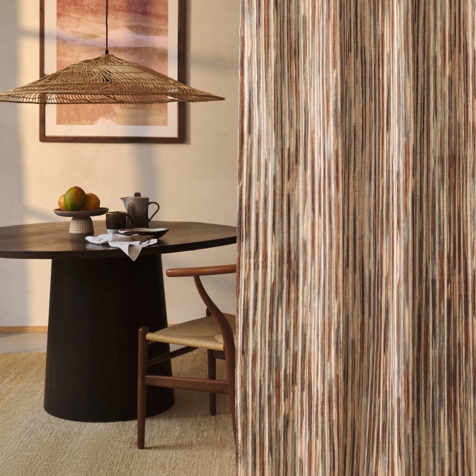Tenda a tutta altezza nel velo Rafi di Villa Nova, colorazione Sienna, accanto a una sala da pranzo con tavolo rotondo scuro, sedie in legno e corda, lampada a sospensione in fibra naturale e quadro alla parete. In vendita c'è soltanto il tessuto al metro: arredi e complementi della scena non sono compresi.