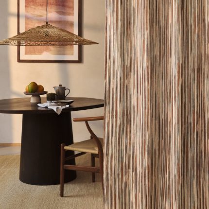 Tenda a tutta altezza nel velo Rafi di Villa Nova, colorazione Sienna, accanto a una sala da pranzo con tavolo rotondo scuro, sedie in legno e corda, lampada a sospensione in fibra naturale e quadro alla parete. In vendita c'è soltanto il tessuto al metro: arredi e complementi della scena non sono compresi.