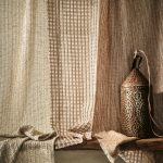 Tenda a tutta altezza nel tessuto Romo Veya colorazione Tawny accanto a un velo chiaro, con una lanterna in metallo traforato e un plaid su una panca in legno. In vendita c’è soltanto il tessuto al metro: arredi e complementi della scena non sono compresi.