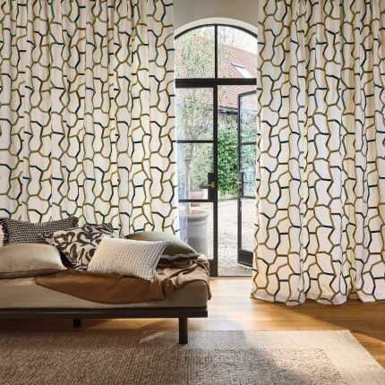 Tende a tutta altezza nel tessuto ricamato Samir di Villa Nova, colorazione Mineral, in una camera da letto con grande vetrata, letto basso in legno scuro e tappeto. In vendita c’è soltanto il tessuto al metro: arredi e complementi della scena non sono compresi.