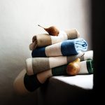 Pezze del tessuto Perry di Élitis piegate e impilate nelle quattro colorazioni tortora, cammello, blu marino e verde bosco, appoggiate su una mensola di pietra con due pere. In vendita è soltanto il tessuto al metro: gli altri oggetti della scena non sono compresi.